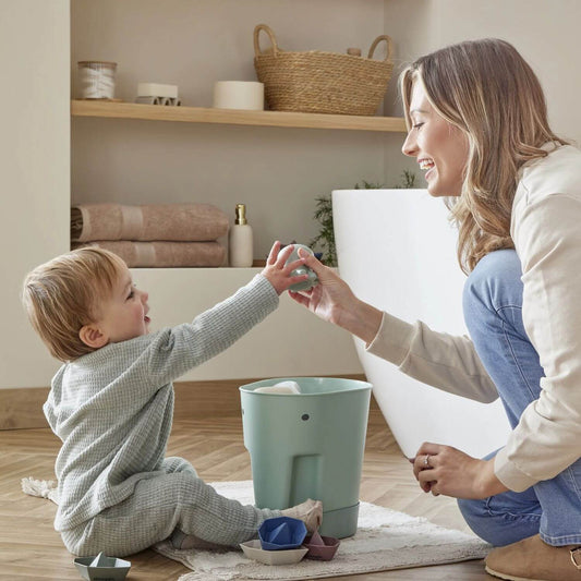 Smiling mother and toddler play with bath toys beside a green Shnuggle drying caddy, in a cosy bathroom with towels and shelves in the background.