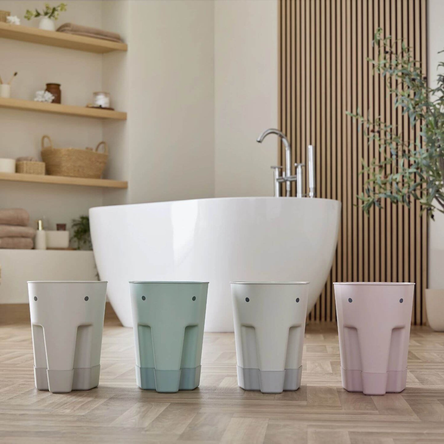 Four Shnuggle bath toy drying caddies in various colours—grey, green, white, and pink—lined up in a modern bathroom with a freestanding tub.