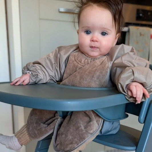 A baby sitting in a highchair wearing a taupe long sleeve towel bib with a soft towelling front and lightweight wipe-clean sleeves, covering the chest and lap during mealtime.