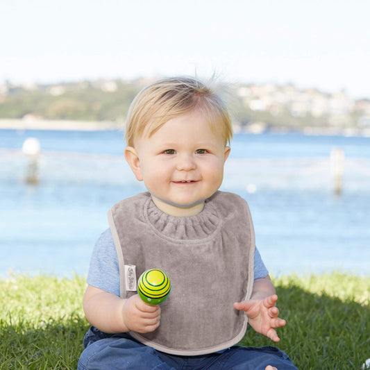 A baby sitting on grass by the water wearing a taupe velour bib, holding a green and yellow toy while smiling outdoors.