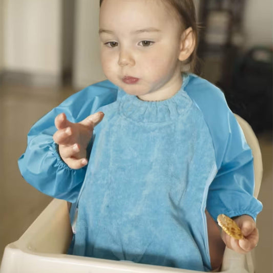 A baby wearing a long sleeve aqua highchair bib while sitting in a highchair, showing full arm coverage, soft towel front and elasticated cuffs during feeding.