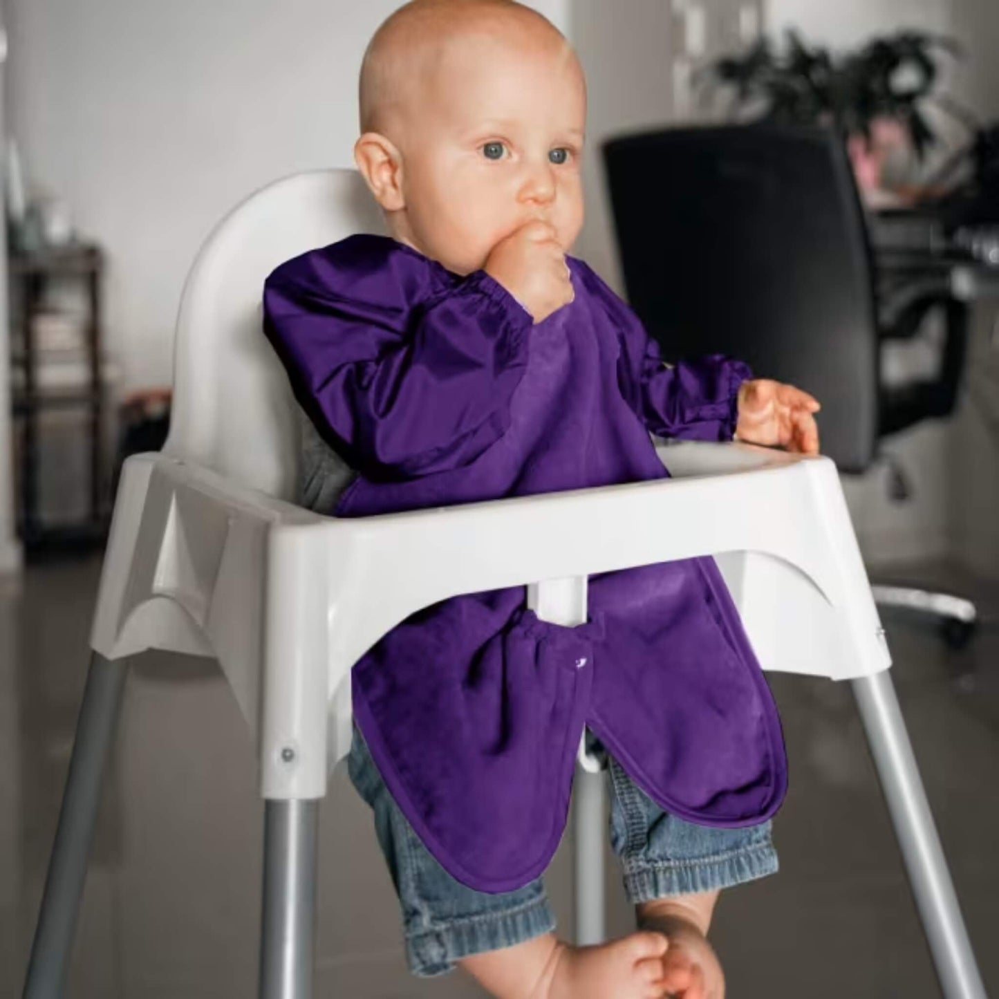 A baby wearing a long sleeve purple highchair bib while sitting in a highchair, showing full arm coverage, soft towel front and elasticated cuffs during feeding.