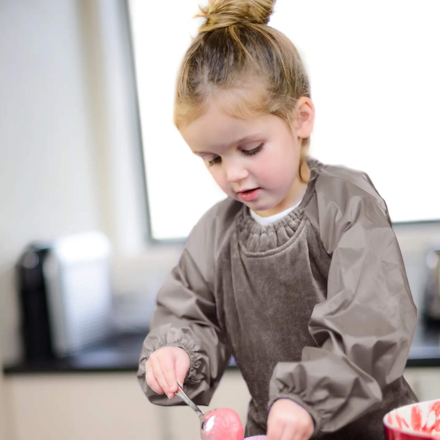 A young child standing at a counter wearing a taupe long-sleeve bib while scooping food from a bowl, with soft natural light in the background.
