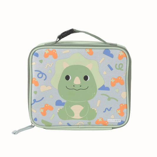 A rectangular kids lunch bag with a top handle, featuring a cute green cartoon dinosaur on a light blue background with colourful retro game controller and confetti patterns. The brand name 'SKIP HOP' is visible on a small tag near the bottom right.