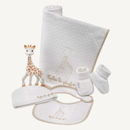 A coordinated newborn gift set laid out flat, showing a white quilted blanket, matching hat, bib, booties, and a natural rubber giraffe toy.