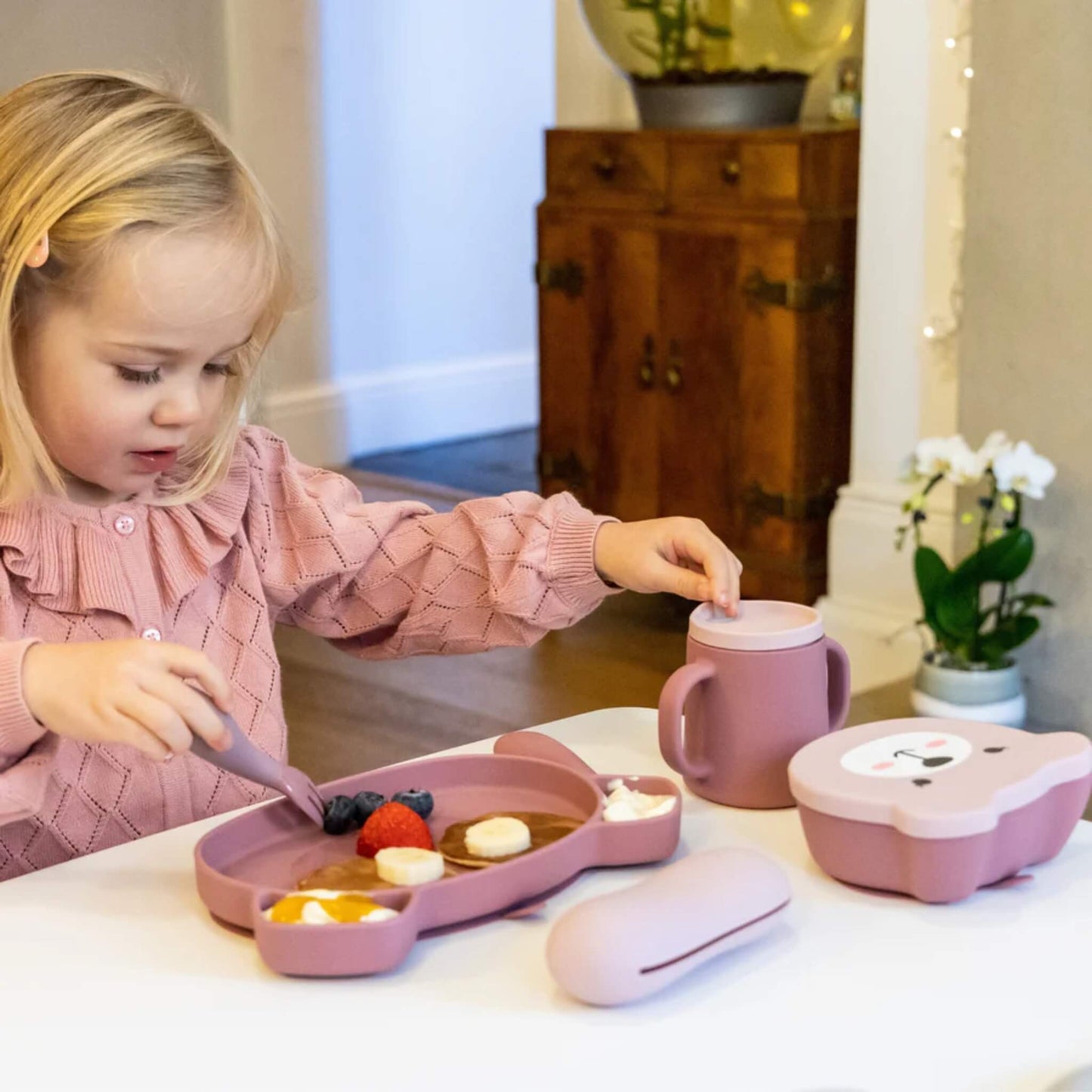 A toddler taking food from a pink silicone plate with a chunky handled fork, with matching bowl and lidded container nearby.