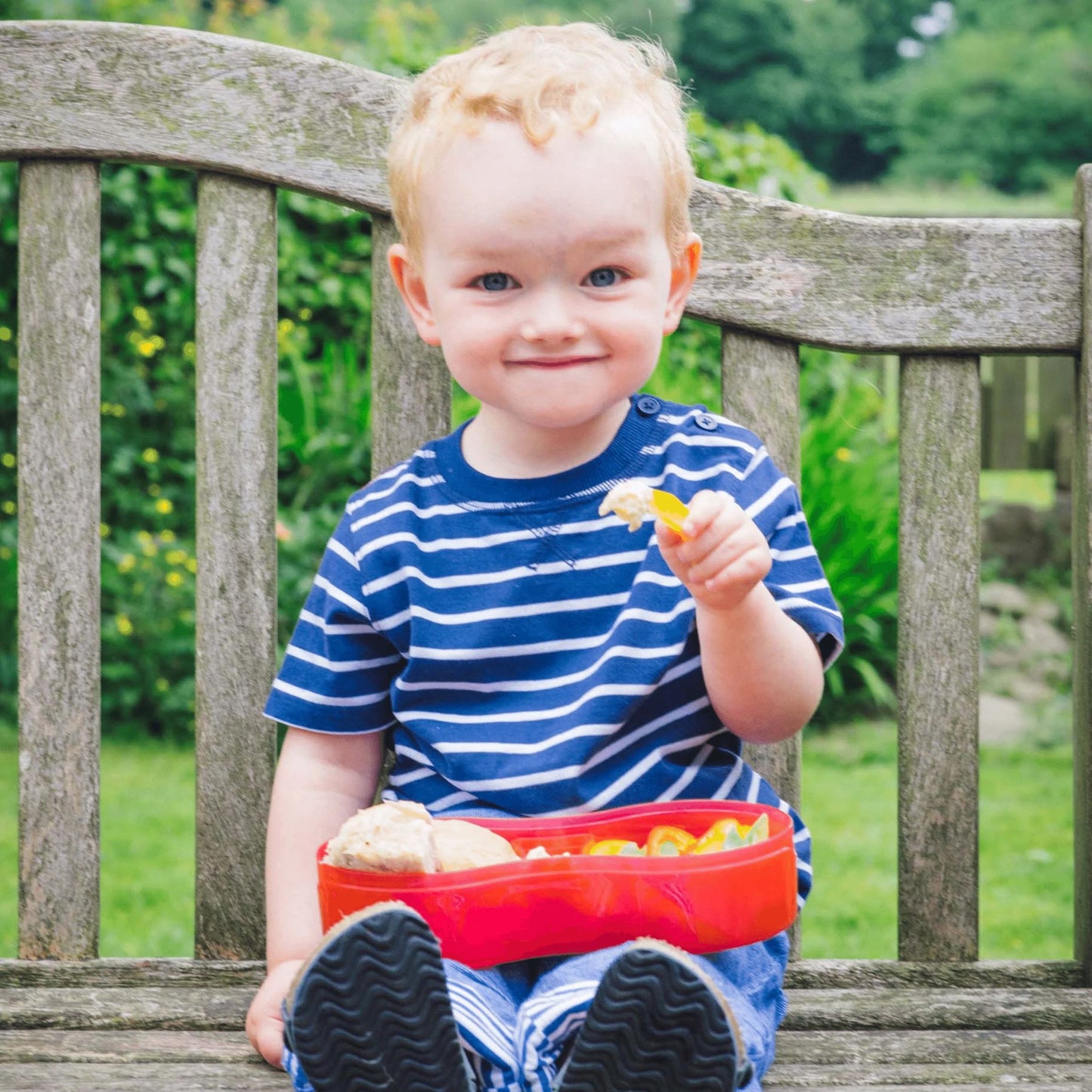 Young child enjoying a meal outdoors with a red lunch box and integrated dipping pot separator, perfect for healthy snacks on the go.