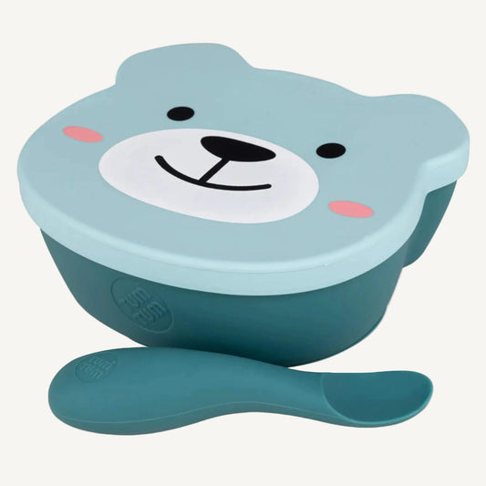 A close-up view of a suction weaning bowl with a fitted lid and a soft weaning spoon placed in front, showing the rounded bowl shape and easy-grip spoon design.
