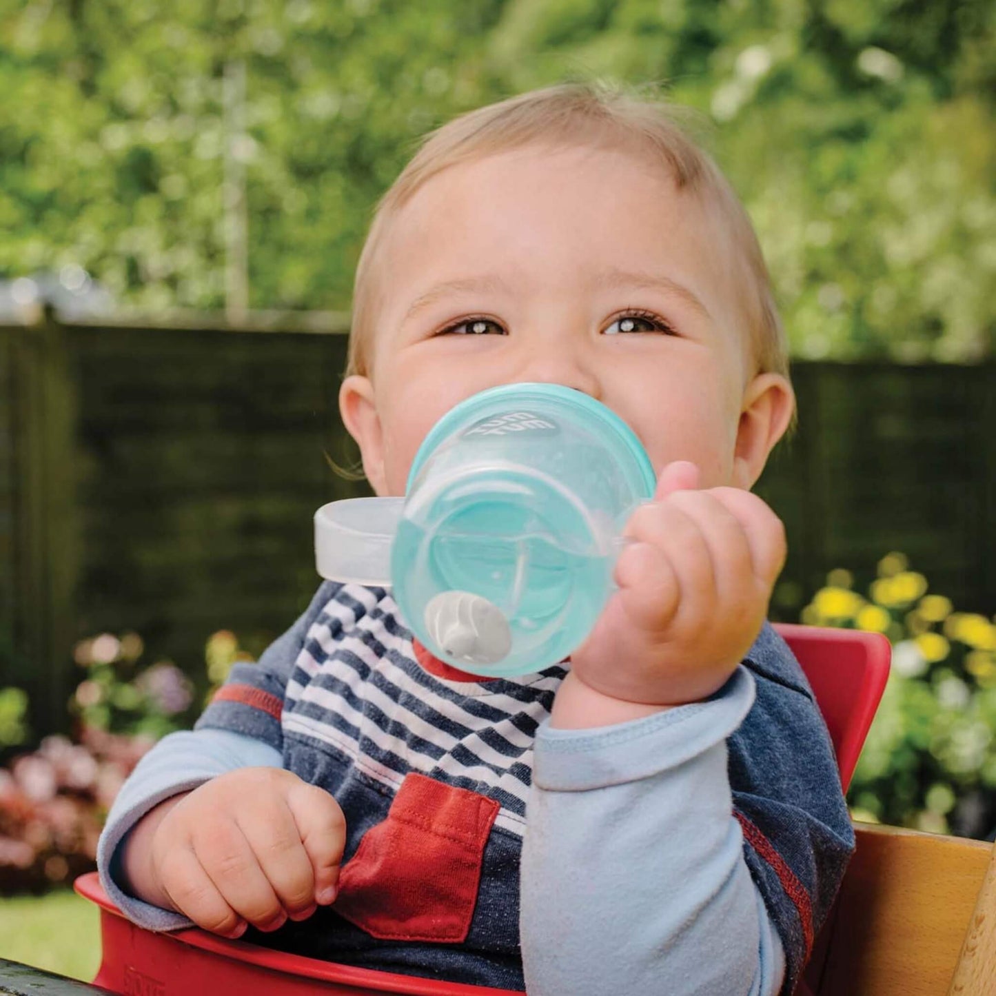 Child holding a blue sippy cup outdoors with greenery in the background
