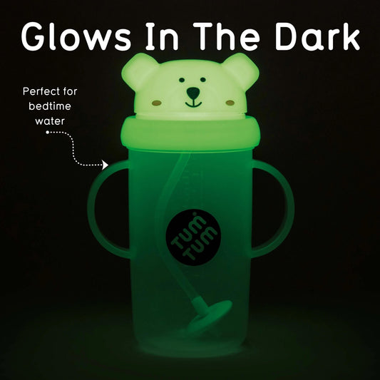 A glow-in-the-dark sippy cup with a bear-shaped lid, shown glowing green in the dark. The cup has two side handles, a straw, and a black circular logo that reads 'tum tum'.
