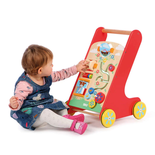 A toddler in a denim dress interacts with the Tidlo Activity Walker, seated beside it and reaching for the spinning activity pieces on the wooden front panel.