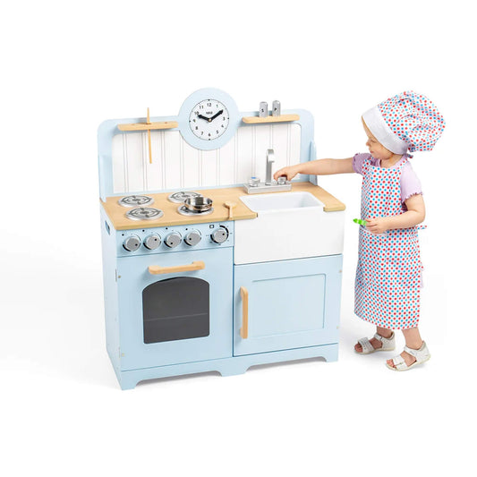 A young child wearing a chef's hat and apron plays at the blue wooden play kitchen, turning the sink tap and holding a spoon.