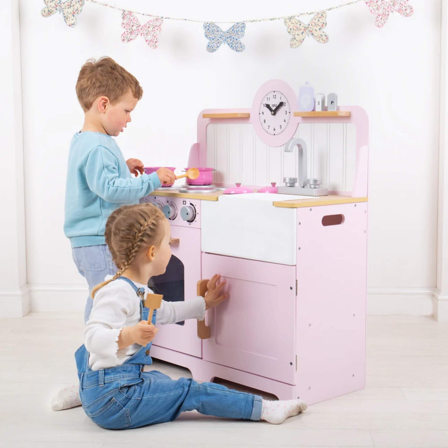 Two children playing with the pink Tidlo wooden play kitchen—one stirring a saucepan on the hob, the other seated and opening the cupboard door below.