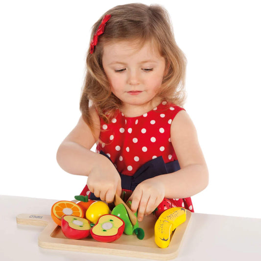 A young girl in a red polka dot dress uses the wooden knife to slice play fruit on the tray, focusing carefully as she cuts through the velcro pieces.