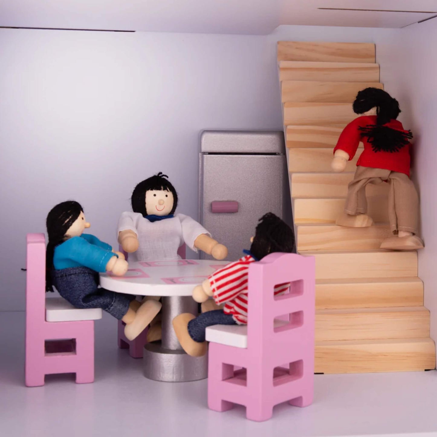 Dolls gathered at a round dining table inside the dollhouse, while one figure climbs the staircase in the background.