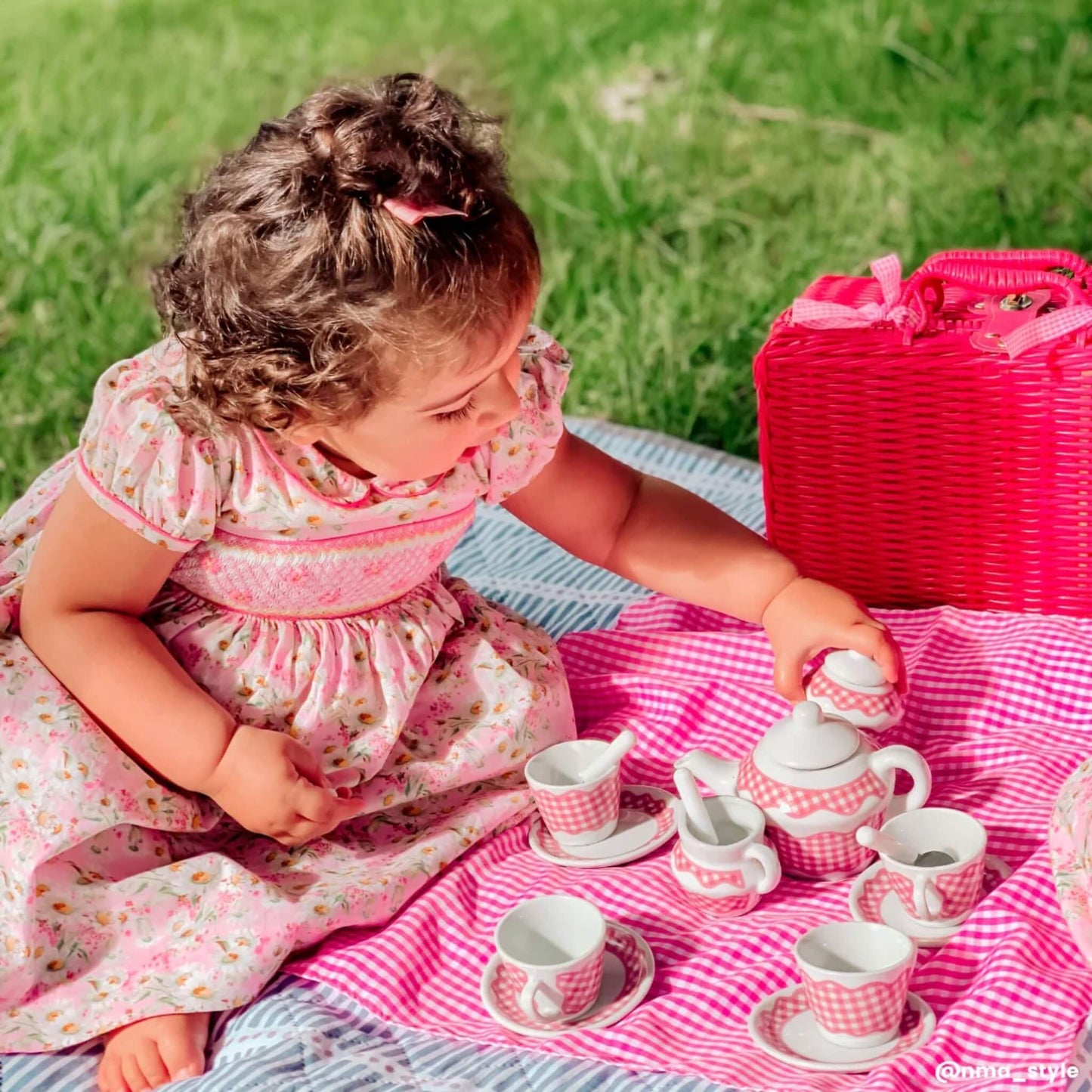 Close-up of a child reaching for the teapot during a pretend picnic, with teacups and saucers laid out neatly on the gingham cloth.