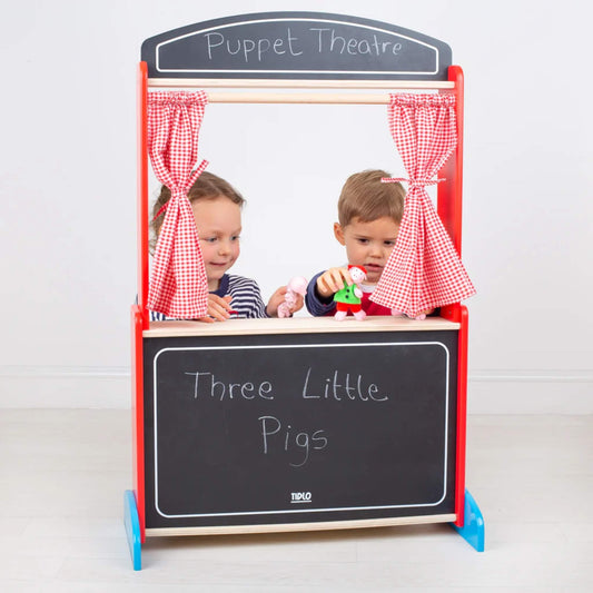 Two children behind a wooden puppet theatre, holding finger puppets on stage with red gingham curtains and ‘Three Little Pigs’ written on the front board.