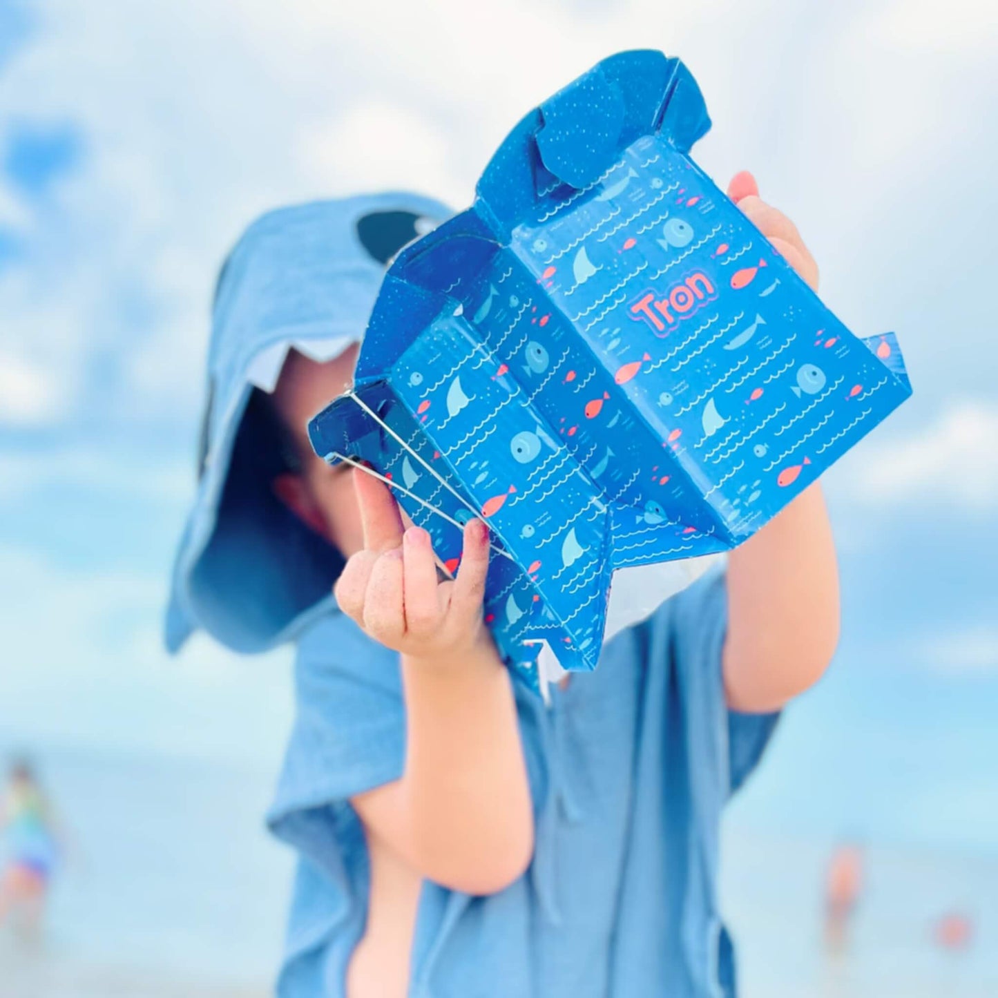 Child in a blue shark hooded towel holds up a folded disposable potty in front of their face against a bright seaside background.