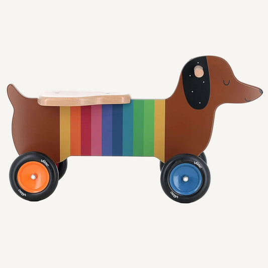 A wooden dachshund-shaped ride-on toy with rainbow stripe body, natural wood seat and four wheels in different bright colours.