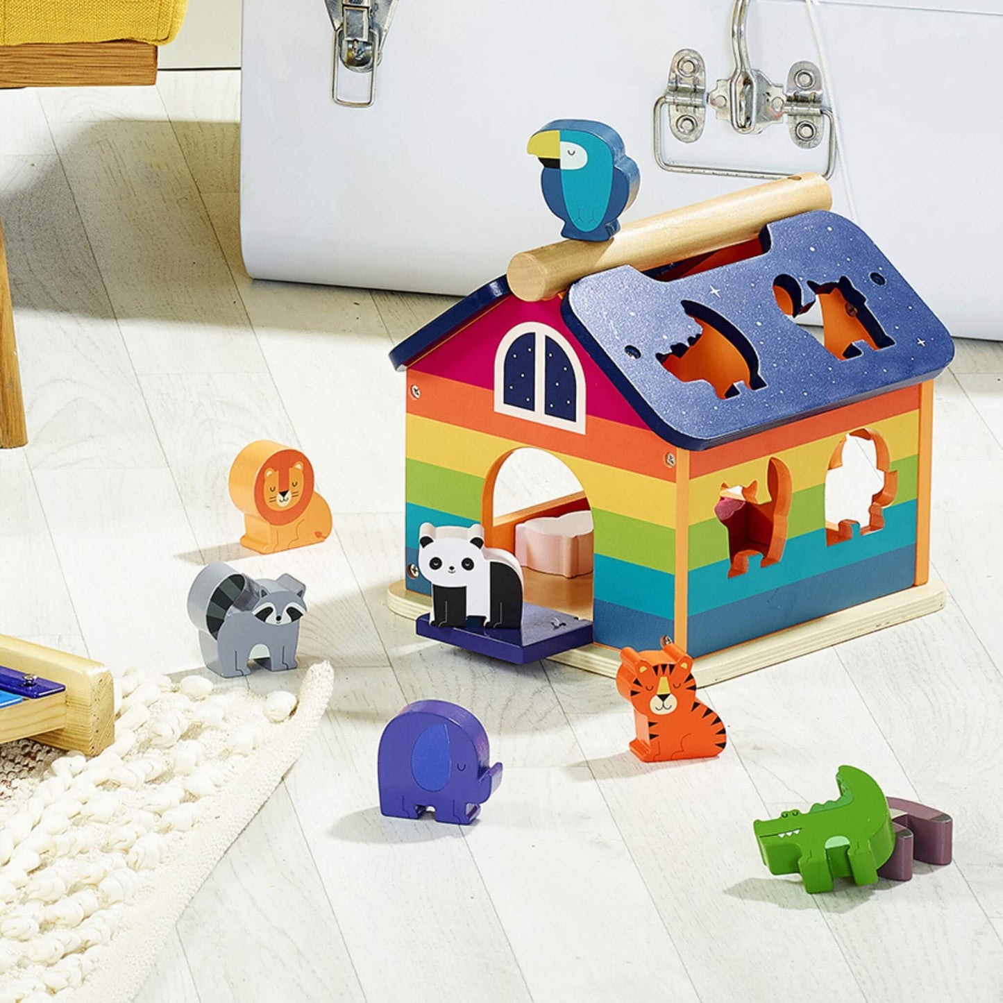 A rainbow-coloured wooden shape sorter with animal blocks positioned on a light wooden floor in a playroom setting.