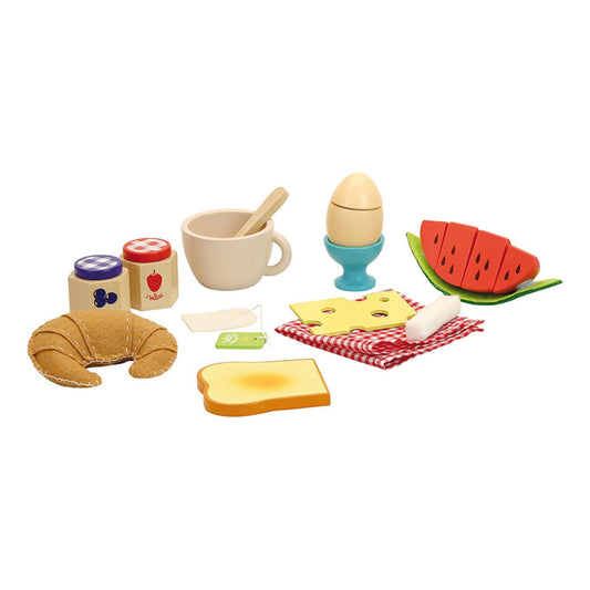 A wooden breakfast toy set arranged on a white surface with croissant, egg in cup, cup with spoon, watermelon slices, toast, cheese slice, jam jars, tea bag, and a small red-check cloth.