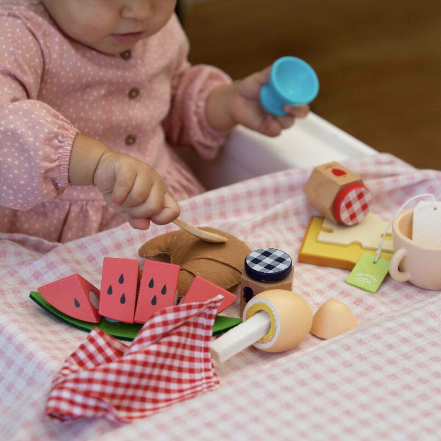 A young child in a pink polka-dot top looking at a wooden egg in an egg cup, surrounded by toy breakfast items including watermelon slices, croissant, jam jars, cheese slice, and toast on a pink-check cloth.