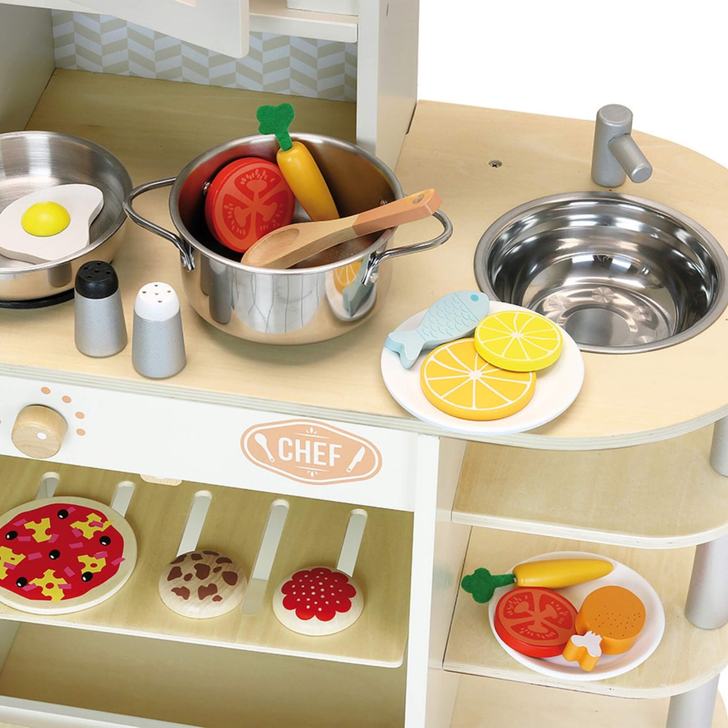 Close-up of a toy kitchen worktop with a saucepan, frying pan, wooden spoon, sink, and play food pieces such as tomato, lemon slices, and fish.
