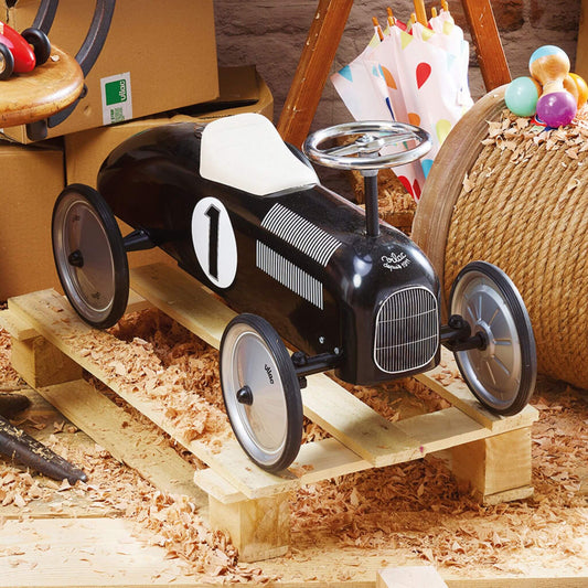 A black metal ride-on toy car with number 1 decal and white seat, displayed indoors on a wooden pallet surrounded by wood shavings and toys.
