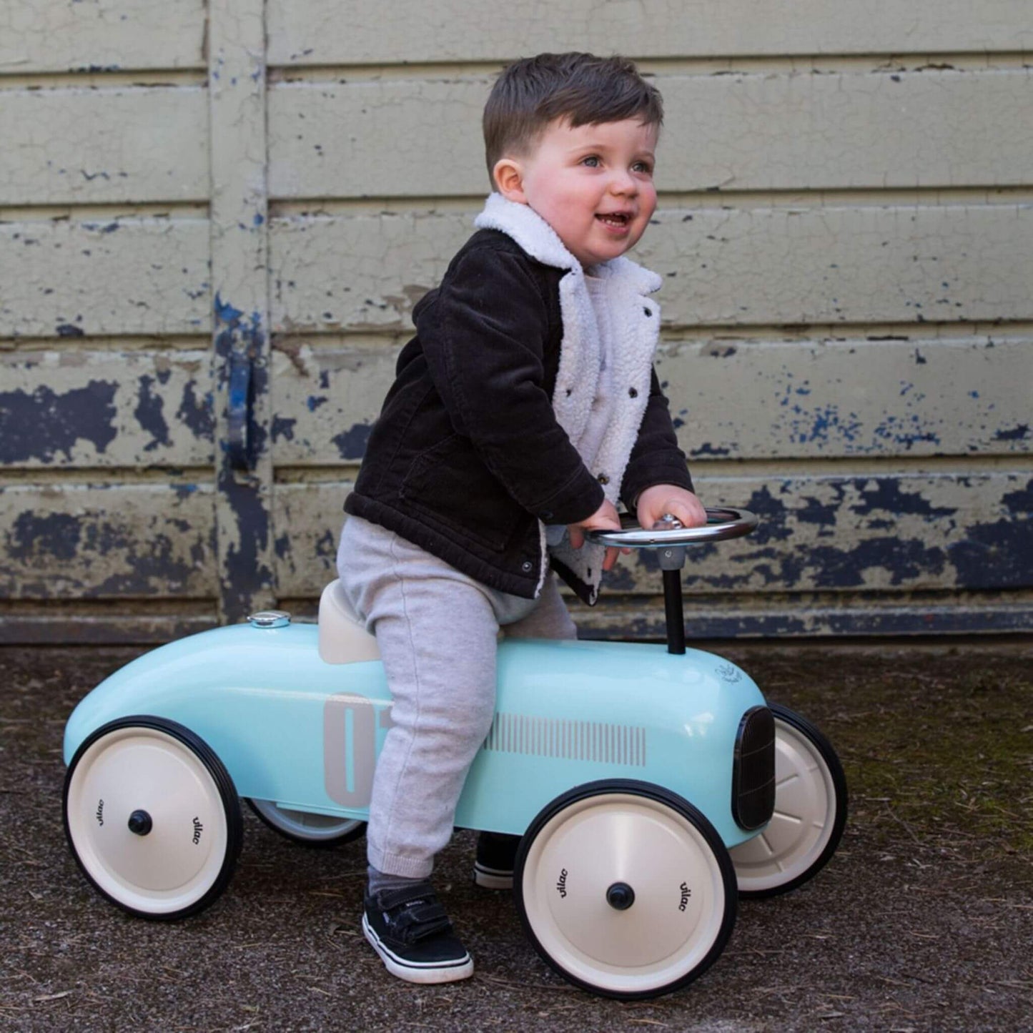 A young child in a grey outfit and black jacket with a white fleece collar is sitting upright on a pale blue metal ride-on toy car, smiling as they look to the side. Both hands are holding the steering wheel, and the car is positioned in front of a worn wooden wall.