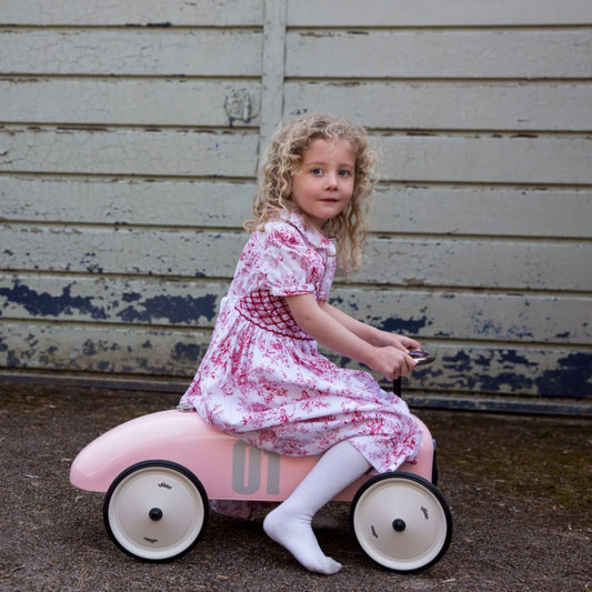 Young girl riding a vintage style metal classic toy car in pink with number 01 detail on the side.