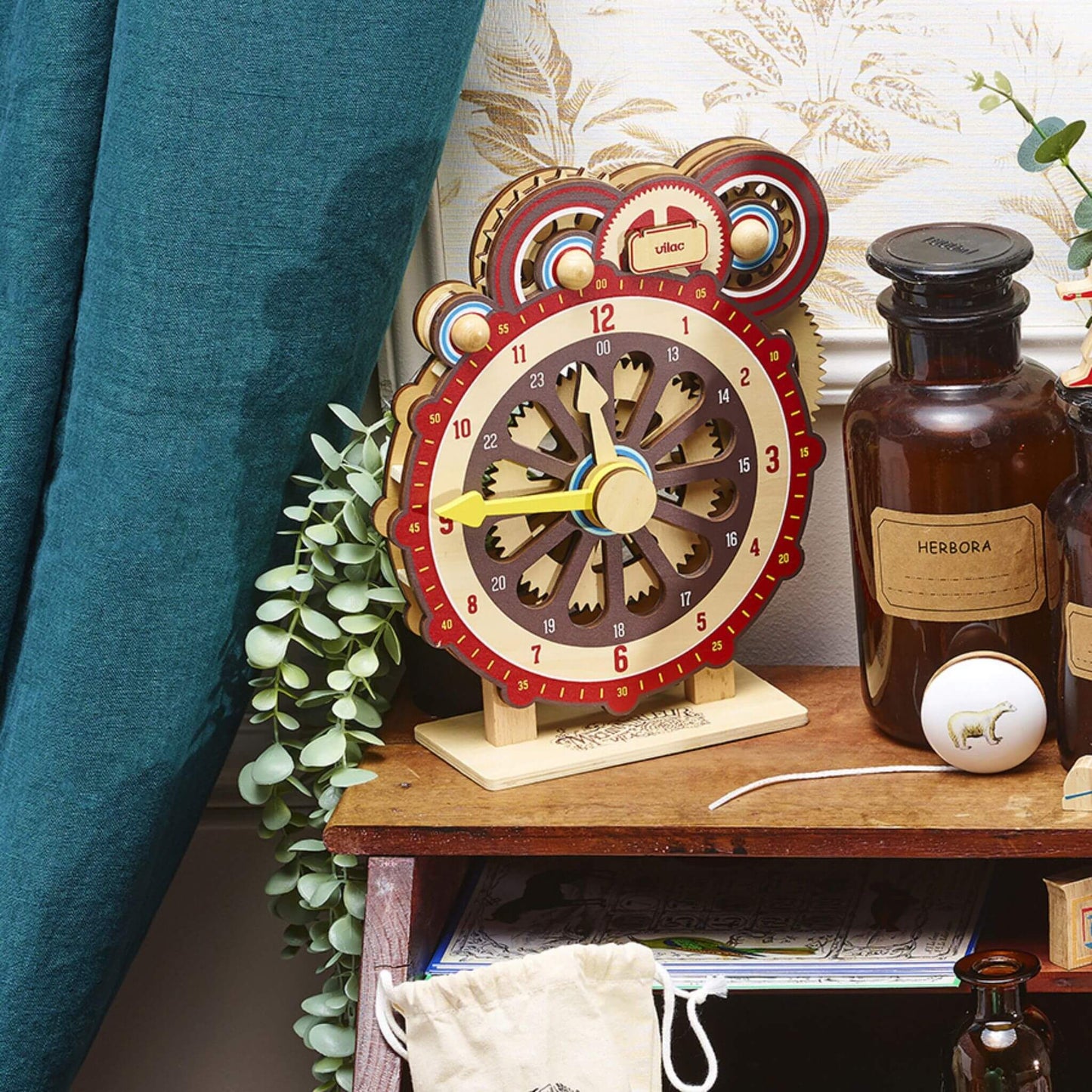 Child's Learning Clock displayed on a wooden shelf with vintage-style bottles, greenery, and a cosy curtain, blending educational play with classic décor.
