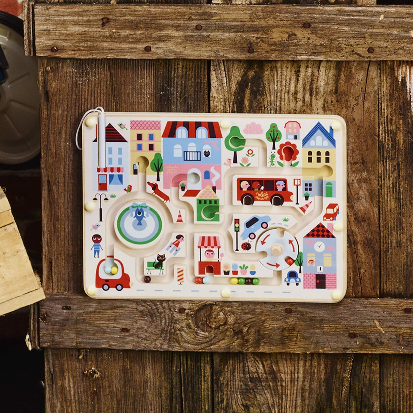Wooden kids town magnetic maze displayed outdoors leaning against wooden planks, showing the full board with houses, cars, bus and fountain details.