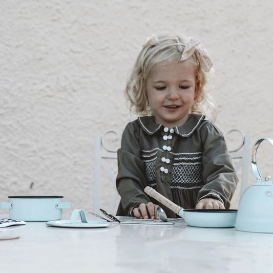 A young girl smiles as she plays with the vintage-style cooking Set, exploring the textures of metal pans and wooden handles.