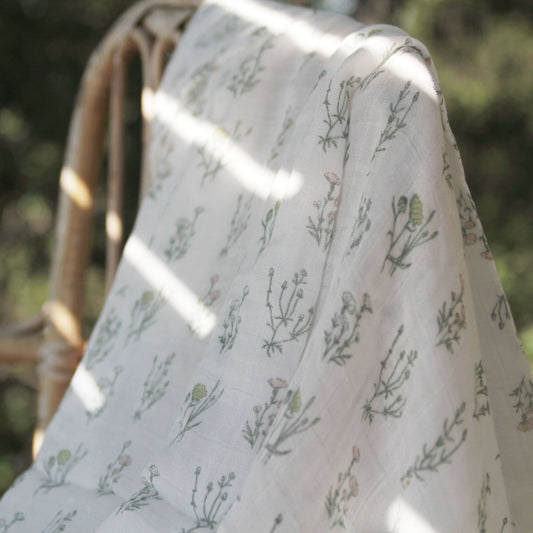 Close-up of aden + anais French Floral Dream Blanket hanging over a chair. Soft white muslin with green and pink botanical details in a hand-drawn style.