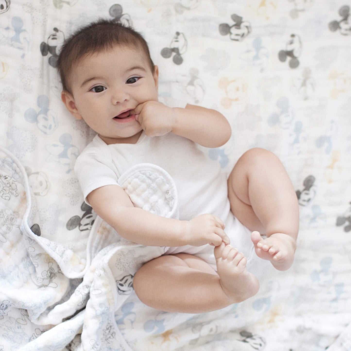 Baby lying on the aden + anais Mickey & Minnie Dream Blanket, surrounded by soft cotton muslin with a Disney character print in calming neutral shades.
