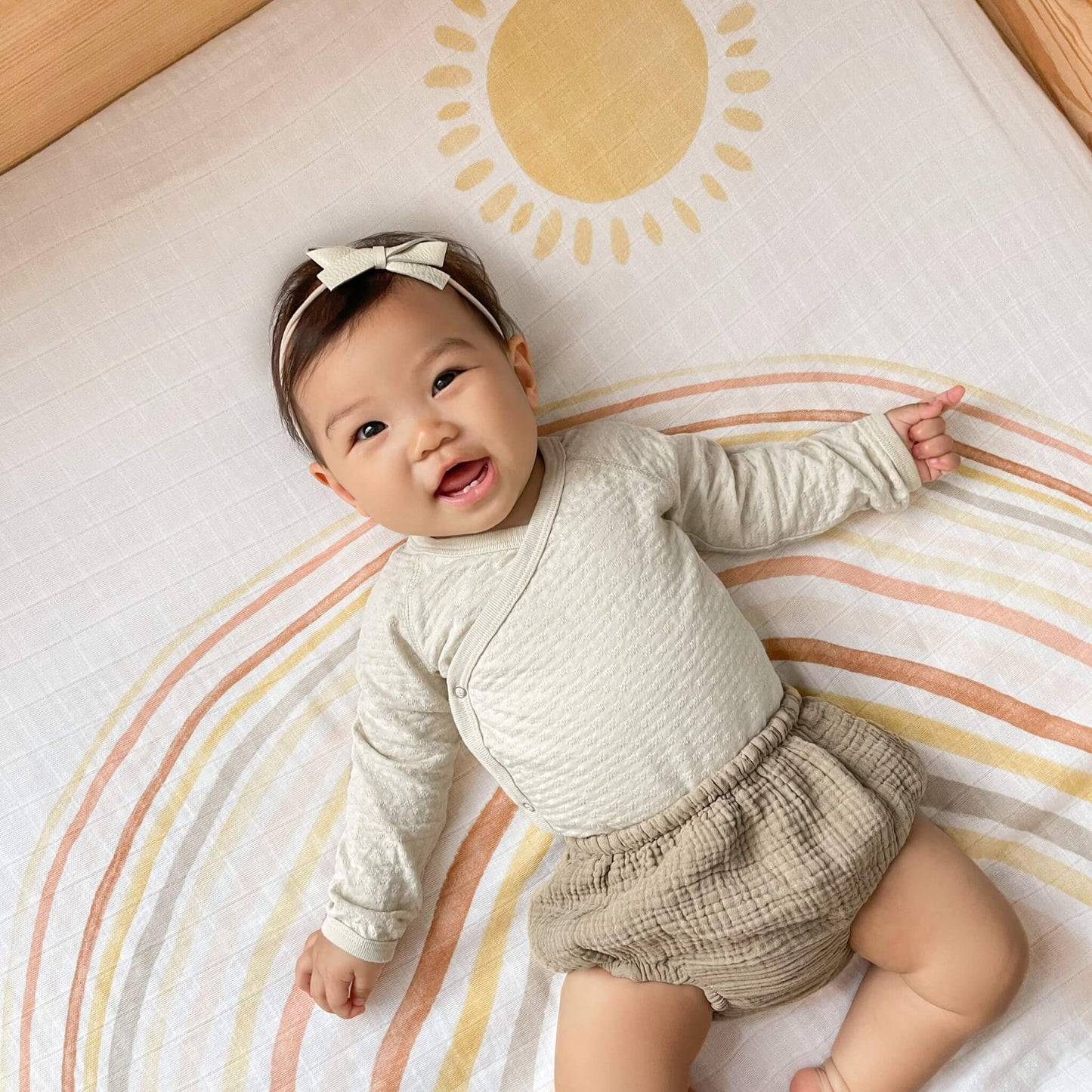 Smiling baby lying on the aden + anais Keep Rising Dream Blanket, made from breathable muslin cotton with a warm-toned rainbow and sun design.