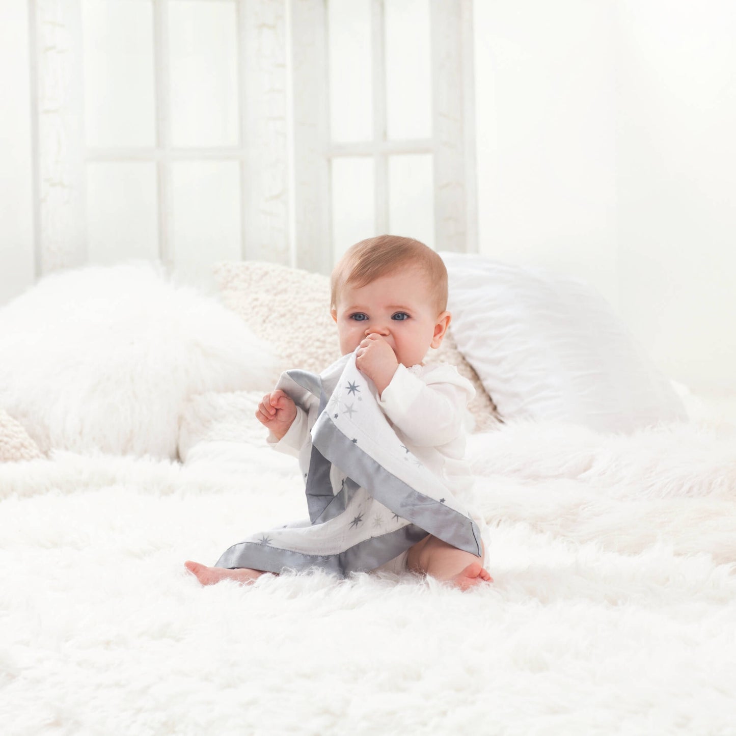 Baby sitting on a fluffy white bed, holding the aden + anais Issie security blanket in Twinkle—soft muslin comforter with satin trim.