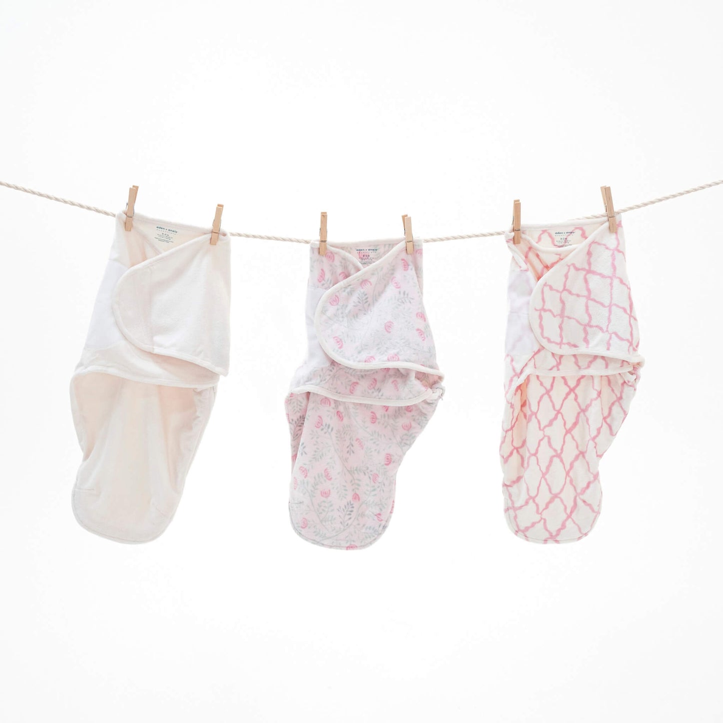 Three aden + anais velboa baby swaddles in Arts & Crafts prints hanging on a clothesline—cream, floral, and pink lattice.