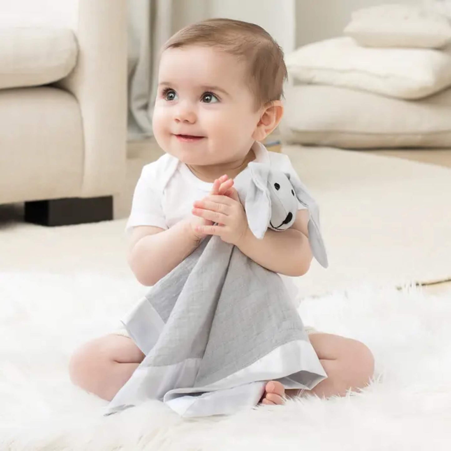 Baby sitting on soft rug holding the aden + anais musy mate lovey in microchip grey. Comforter made from cotton muslin with satin trim and cuddly plush head for soothing.