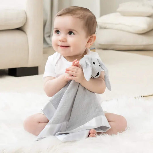 Baby sitting on soft rug holding the aden + anais musy mate lovey in microchip grey. Comforter made from cotton muslin with satin trim and cuddly plush head for soothing.