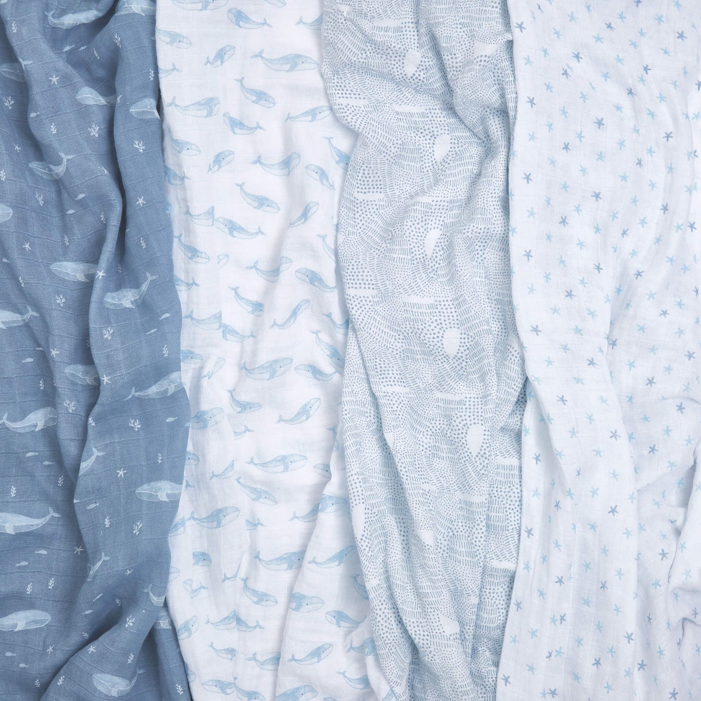 Flat lay of four crumpled Oceanic muslin swaddles in soft blue prints. Features whales, starfish, and ocean-inspired textures on organic cotton.