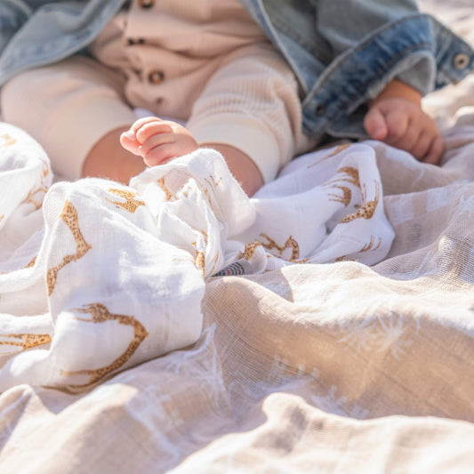 Close-up of baby feet resting on aden + anais Safari Dreams swaddles with giraffe and tree prints in a sunlit outdoor setting.
