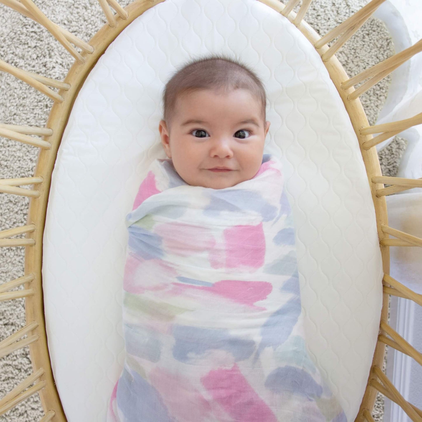Baby swaddled in a silky soft aden + anais bamboo muslin blanket, resting in a woven bassinet.