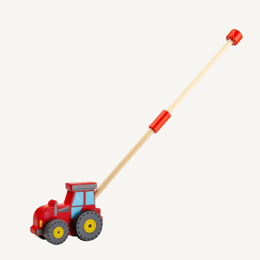 Red wooden tractor push along toy with a long natural wood handle extended behind it, featuring chunky grey wheels, yellow wheel centres and painted cab windows.