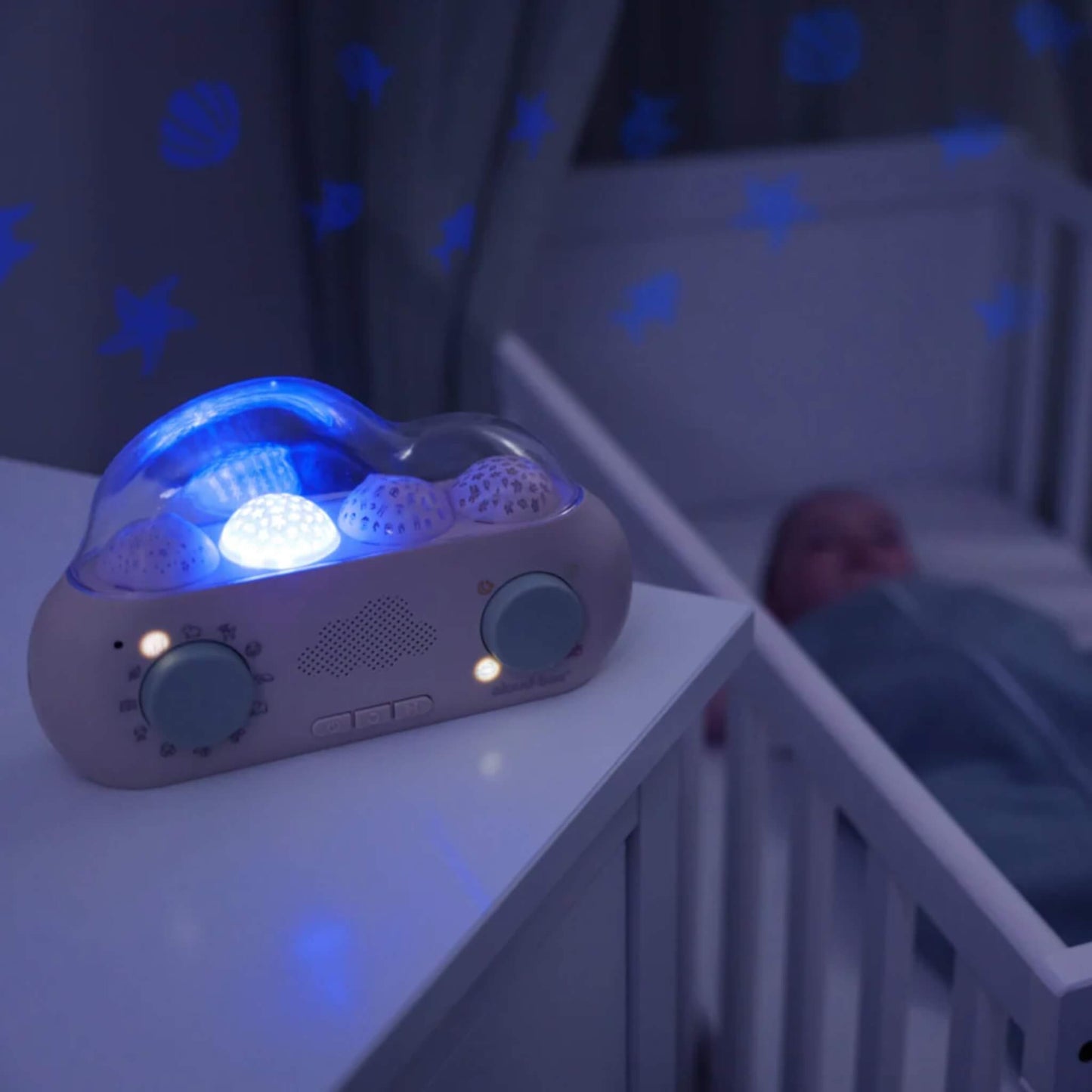 A CloudBox glowing blue on a bedside table and projecting blue stars and other shapes onto the wall beside a sleeping baby in a cot.