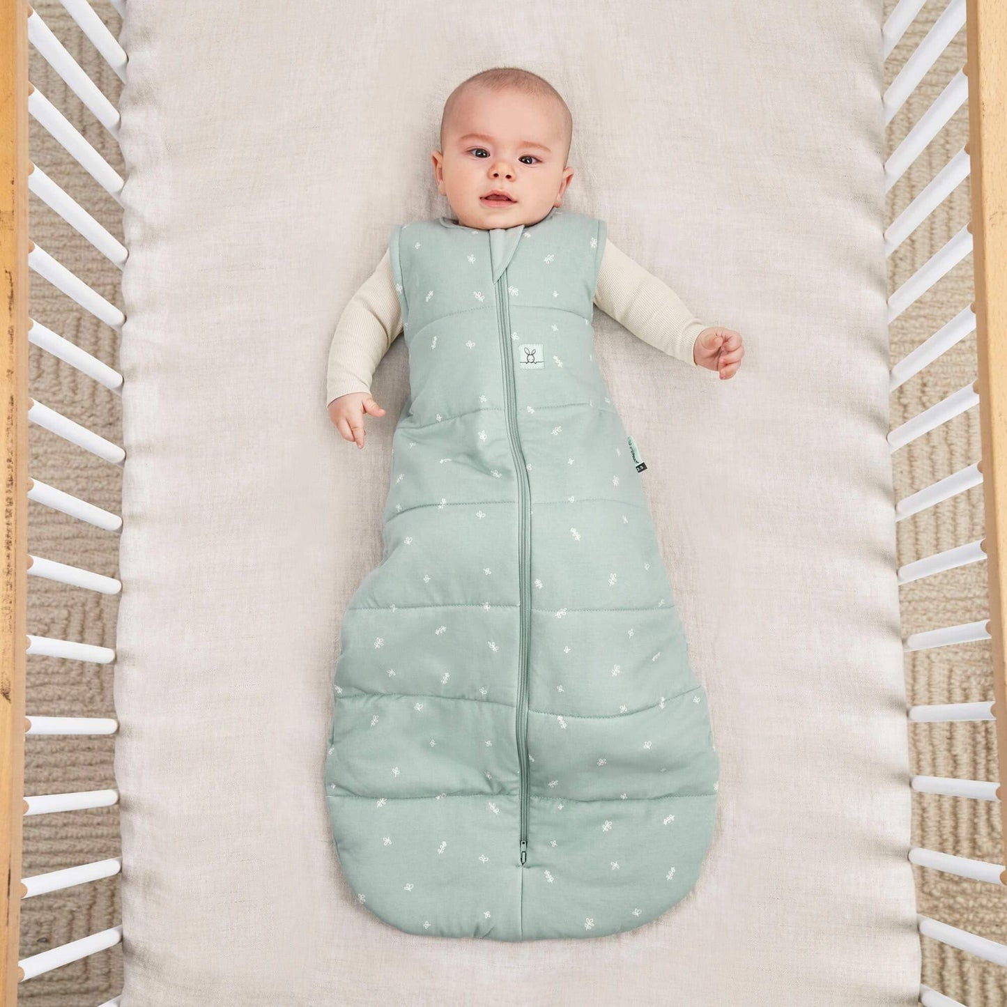 A baby lies in a cot wearing a sage green winter weight sleeping bag, with a central zip and small white patterns.