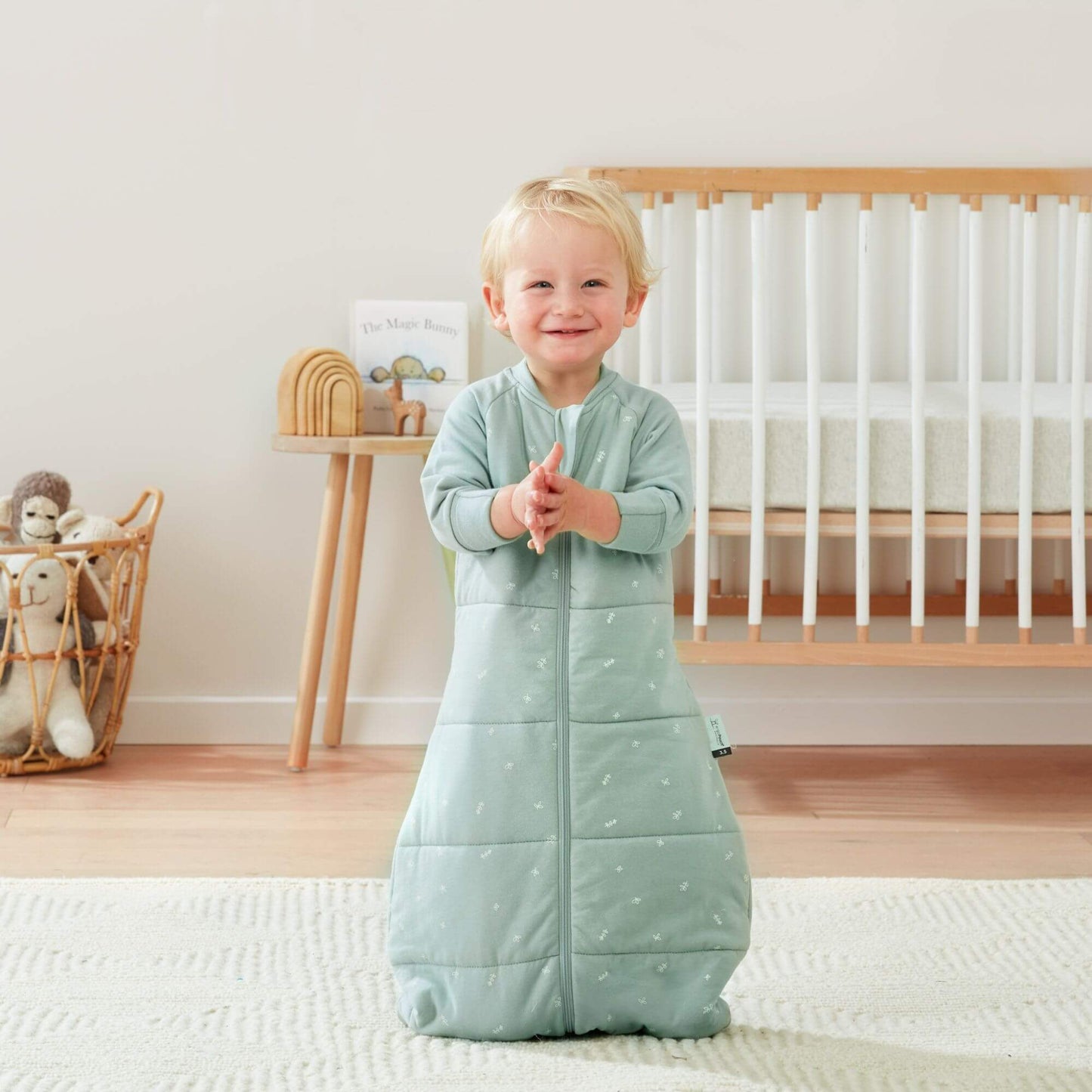 A smiling toddler claps while wearing a sage green sleeping bag, in a nursery with a cot and toys.