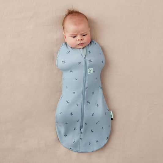 Baby lying on a neutral surface swaddled in a light blue Cocoon Swaddle Bag decorated with dragonfly print, both arms tucked inside.