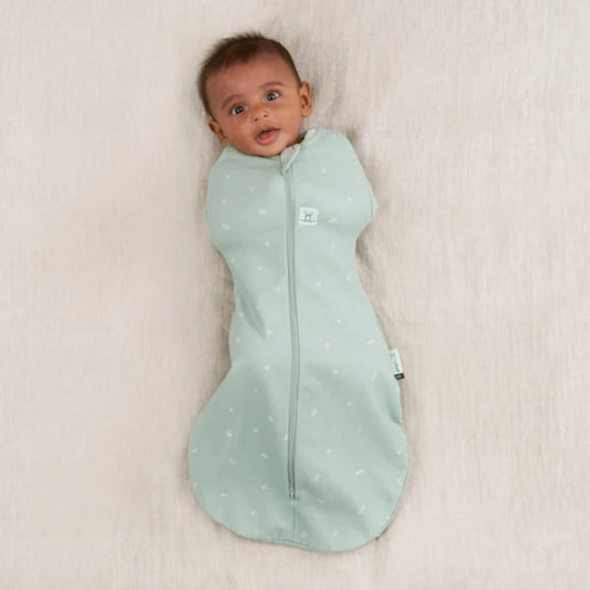 Baby lying on a neutral blanket wearing a sage green newborn swaddle bag with zip closure and arms gently secured inside.