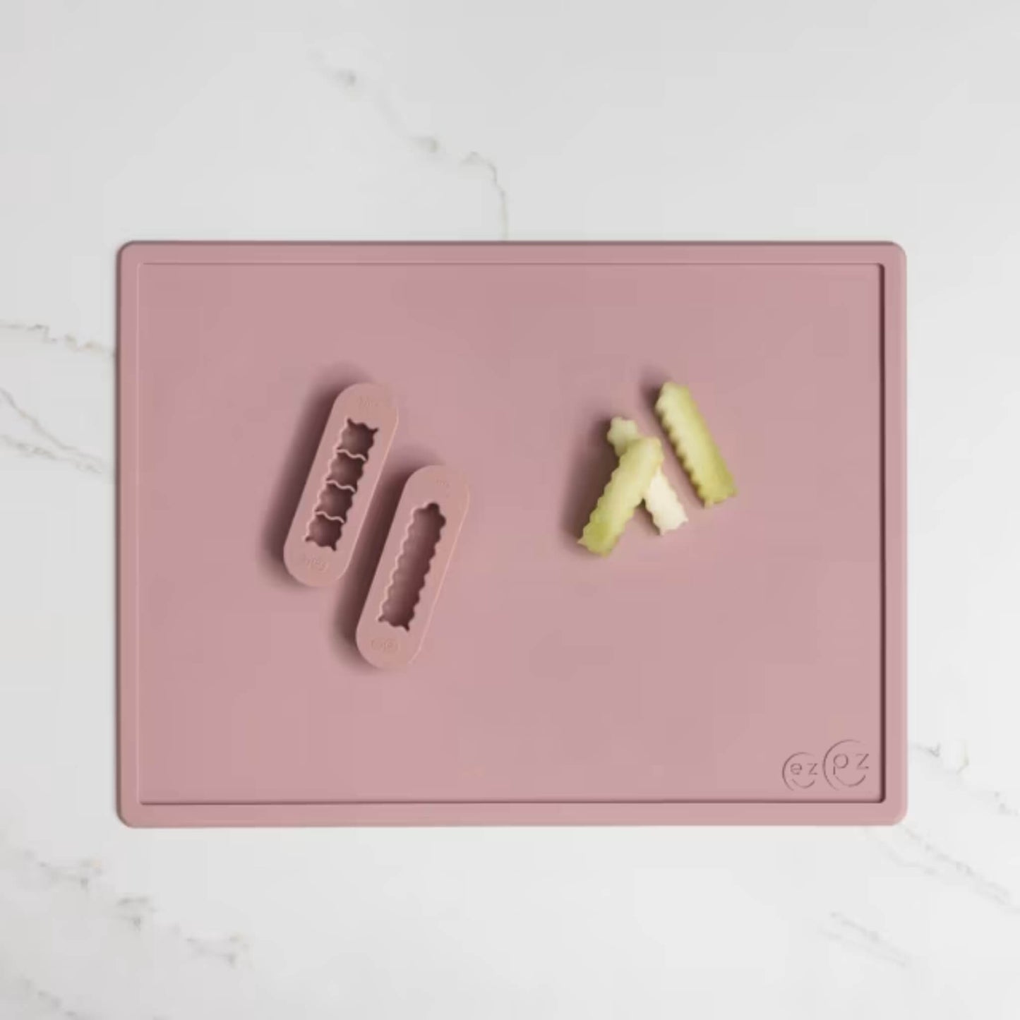 Blush pink silicone food cutters beside small pieces of soft fruit prepared into baby-friendly strips on a matching placemat.